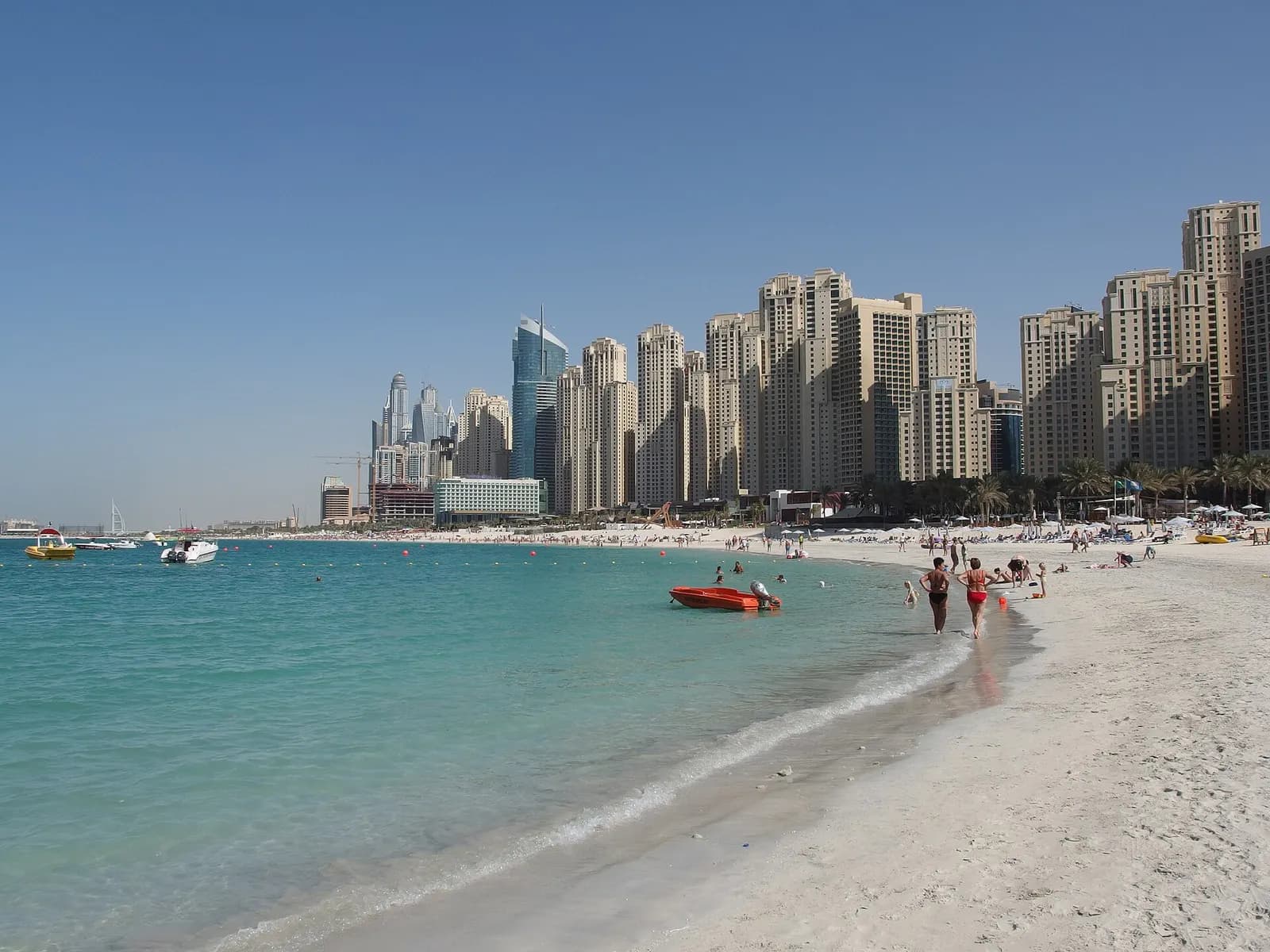 Daytime view along the JBR beach, with the sea on the left and a row of residential towers behind the sand, and the Dubai Marina skyline in the distance.