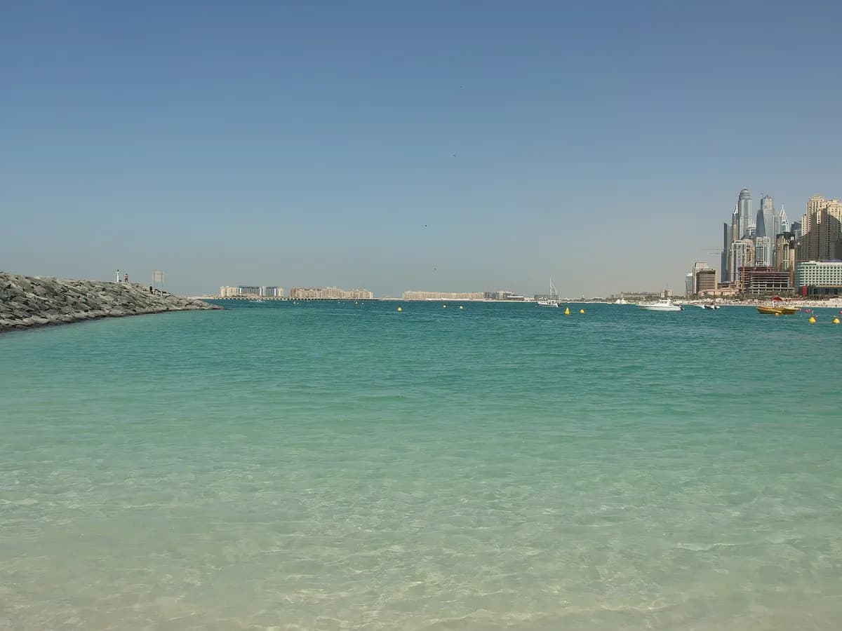 Calm shallow water at the JBR shoreline on a clear day, with a rock breakwater on the left and the Dubai Marina and JBR skyline at the right edge.