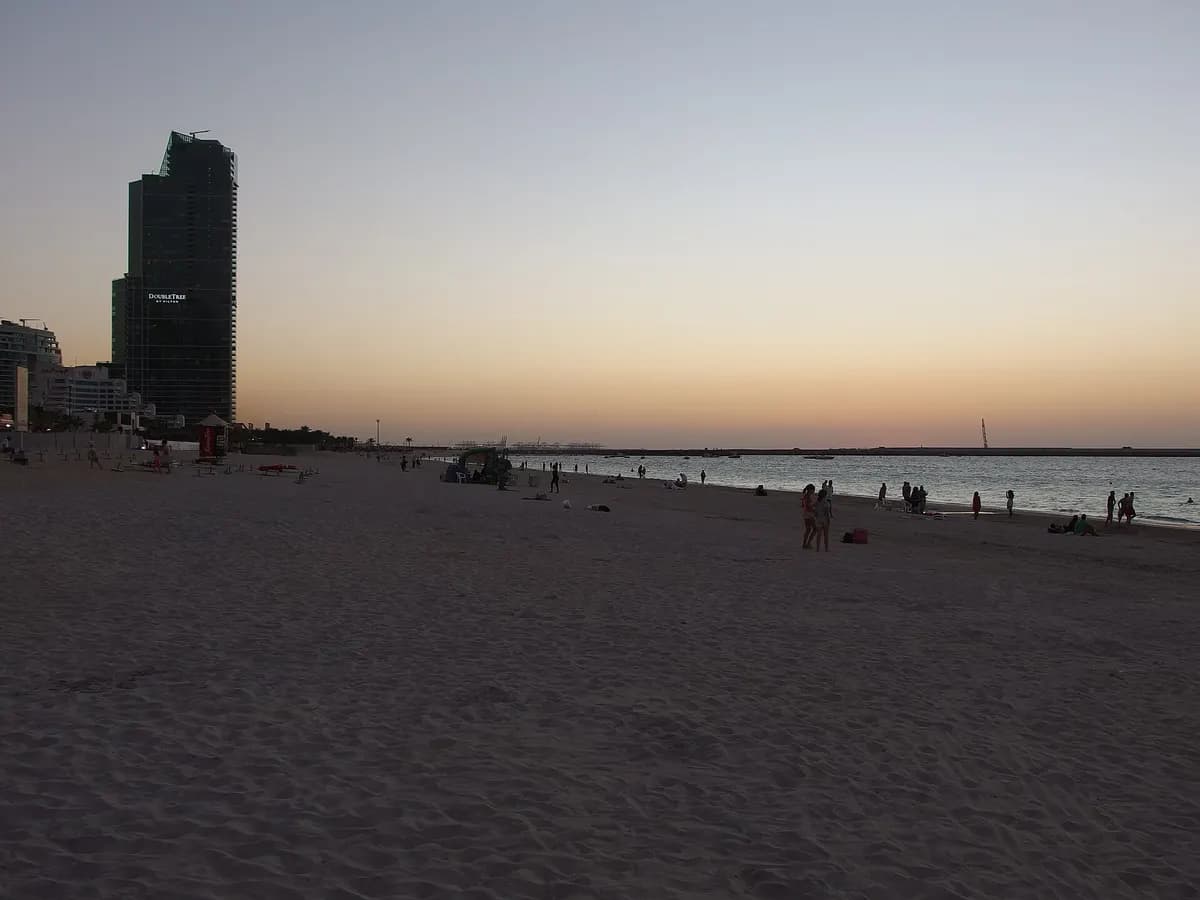 Wide sandy beach at dusk, with the DoubleTree by Hilton tower on the left and people along the water on the right.