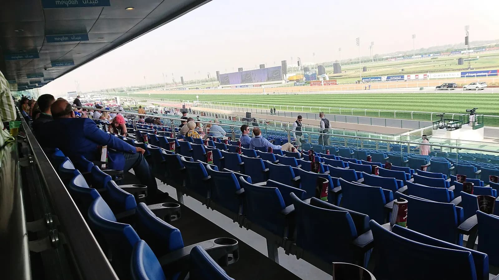 Daytime view from the covered stands at Meydan Racecourse, over rows of blue seats to the striped turf track and the dirt track beyond.