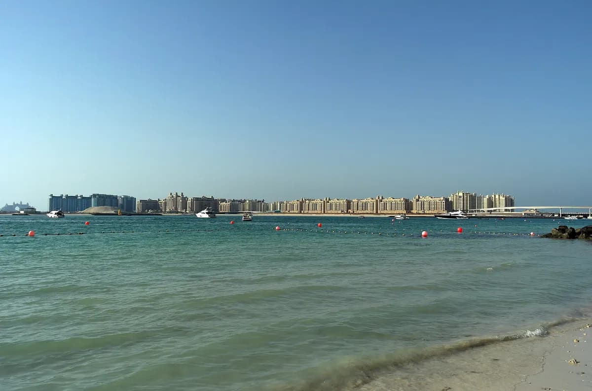 Rows of sand-coloured apartment buildings along the shore of Palm Jumeirah, seen across the water from a beach on a clear day.