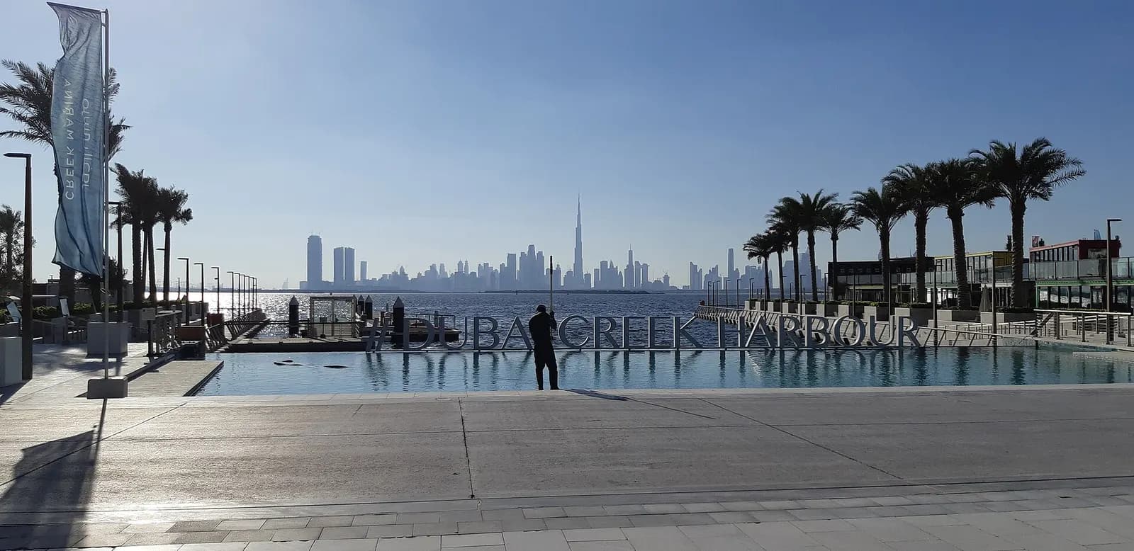 The Dubai Creek Harbour name in large letters over a waterfront pool, with palm trees on both sides and the Dubai skyline in the distance across the water.