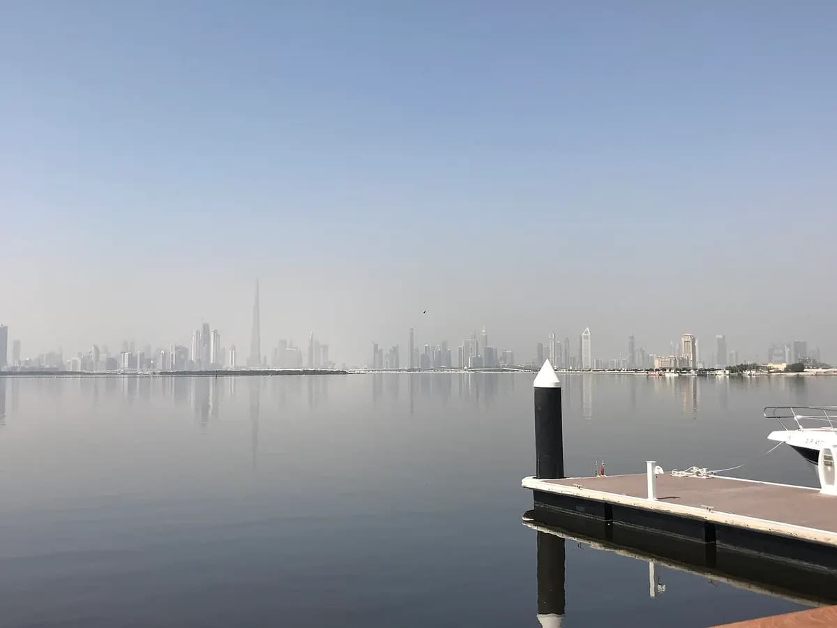 A floating dock with a moored boat on calm water at Dubai Creek Harbour, with the Dubai skyline and the Burj Khalifa far across the creek in haze.