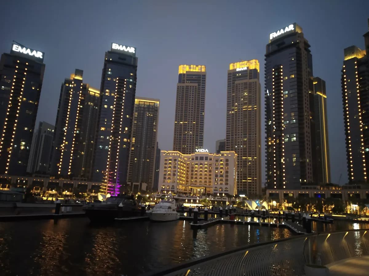 Lit residential towers and a low hotel building behind a marina with moored boats at Dubai Creek Harbour at dusk.